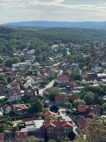 A wide aerial view shows a European village with red-roofed houses nestled among rolling green hills. The camera pans down, focusing on a winding road that meanders through the village. Several cars can be seen driving on the road and parked in driveways. The architecture of the houses is a mix of traditional and modern styles, with many featuring white walls and dark wooden beams. Lush green trees and gardens surround the houses, creating a picturesque and tranquil scene. The sky is partly cloudy, with the sun occasionally breaking through to illuminate the village.