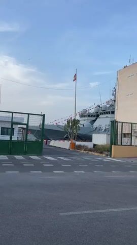 The video opens with a shot of a paved area in front of a gate. In the background, two gray warships are docked. A Spanish flag flies from a flagpole on the right. A single cyclist wearing a helmet and a bright orange vest rides across the paved area from left to right. The cyclist is the only person visible. The weather appears to be clear with a partly cloudy sky. The area looks like a port or naval base.