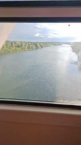 The video shows a view from inside a vehicle, likely a train or bus, looking out a window. The view is of a wide river with green trees and vegetation lining the banks. The sky is blue with some clouds and the sun is shining, creating a glare on the window. The camera pans slightly as the vehicle moves, showing a continuous view of the river and its surroundings. There are no people or specific objects clearly visible outside the window. The interior of the vehicle is only visible as the edges of the window frame and a part of the seat or dashboard.