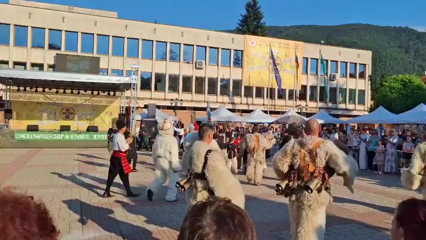 The video shows an outdoor festival or event taking place in a public square in front of a modern, multi-story building with a flat roof. The building has a facade with numerous windows and is adorned with flags and a large banner. The area is set up with several white tents, suggesting vendor stalls or event areas. A stage or platform is visible on the left side of the frame. In the center of the square, a group of people are performing or parading. Many of them are wearing elaborate, furry costumes, some adorned with bells, suggesting a traditional folk performance, possibly related to a carnival or festival. These performers are in motion, with some dancing or walking. The background shows trees and a hillside, indicating a possibly mountainous or hilly terrain surrounding the town. A crowd of spectators is gathered, observing the performance. The sky is clear and blue, suggesting fair weather, and the lighting indicates it is daytime, likely late afternoon due to the angle of the sun.