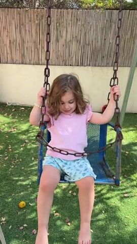 A young girl with curly brown hair, wearing a pink t-shirt with ruffled sleeves and light blue shorts with a cloud pattern, sits on a blue swing set. She is holding onto the chains of the swing with both hands and looking down. The swing is suspended from metal chains. The background consists of a bamboo fence and green artificial turf. A yellow fruit and a red ball are visible on the turf. The girl appears to be in the process of swinging, as evidenced by the slight forward lean and the movement captured in the frames.