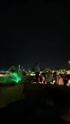 A nighttime video shows a public area with a fountain. The fountain has green and blue lights illuminating the water. Several people are standing near the fountain, some are taking pictures. One person is standing on a ledge to the left of the fountain, and others are gathered in a group to the right. The background shows more lights and some trees. The sky is dark and clear.