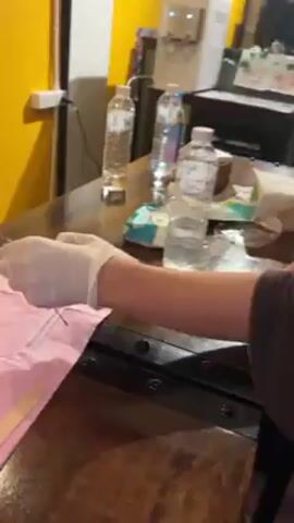 A young woman wearing white gloves is preparing a needle-like object at a wooden table in a room with a yellow wall. She looks up directly into the camera. Bottles of water, wrapped disposable wipes, and other items are on the table. A water dispenser is visible behind her. The woman has brown, wavy hair and wears a brown off-shoulder top.