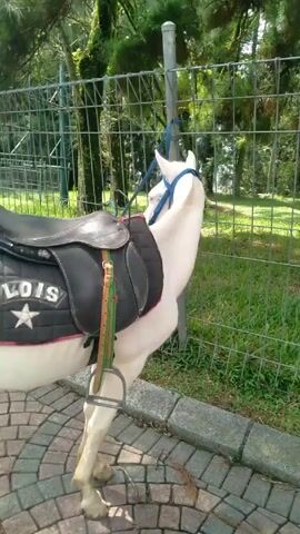 The camera is focused on the rear of a white horse with a black saddle labeled “LOIS” and a star on the side, tied to a metal post at the edge of a brick-paved area. In the background, along the path, are men riding horses in the same direction away from the camera. The area has green grass, a fence, and trees.