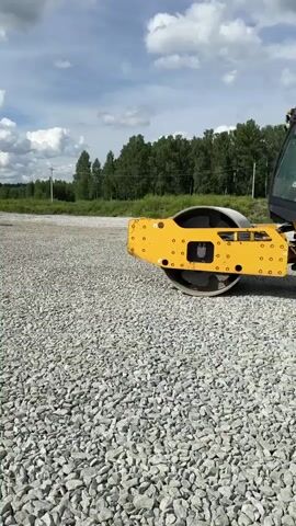 A yellow road roller is being operated on a gravel surface under a partly cloudy sky. The roller moves from left to right across the frame, its large front drum and rear tire visible. The landscape behind the roller features a line of trees and vegetation, with some power lines visible in the distance. The sun is casting shadows, indicating it is daytime. The video is shot from a low angle, close to the ground.