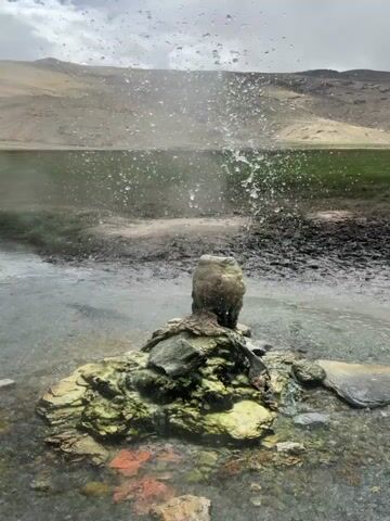 A geyser erupts in a natural landscape. The geyser consists of a cone-shaped mound of rock and minerals with greenish and reddish coloration. Water forcefully sprays upwards from the center of the mound, creating a plume of mist and droplets. The geyser is situated in a shallow body of water with a muddy bank to its right. In the background, there is a green, grassy field that transitions into a barren, tan-colored hillside under a cloudy sky. A person's hand is visible on the right side of the frame, extended towards the geyser. The video captures the dynamic eruption of the geyser, with water splashing and falling around the mound.