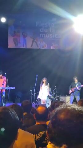 A female singer in a white dress performs on a stage in front of an audience. She has long, dark curly hair and is wearing white knee-high boots. She is holding a microphone and dancing. To her left, a keyboardist in a light-colored shirt plays a keyboard. To her right, a guitarist in a dark shirt plays a guitar. Behind her, a drummer is visible. Above the stage, a large banner displays "Fiesta de la Música" along with illustrations of people playing instruments and dancing. The stage is lit with blue and white lights, creating a smoky atmosphere. The audience is visible in the foreground, with their backs to the camera.