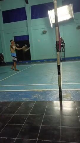 The video shows a badminton court with a net set up. The walls are painted blue and have some blue and green fabric draped across them. There are two men playing badminton. One man is wearing a blue shirt and shorts, and the other is wearing a red shirt and black shorts. The first man is holding a badminton racket and appears to be in the middle of a game. The second man is also holding a racket and is positioned on the opposite side of the net. The lighting in the court is from overhead fluorescent lights. The floor is tiled with black and white squares. There is also a fan visible on the wall in the background.