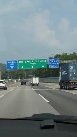 The video shows a view from inside a vehicle traveling on a highway in South Korea. Ahead, there are several signs indicating directions and distances. One sign, in green, reads "현금,전자카드 요금내는곳 Tollgate 1.0 km" with two downward arrows. Another blue sign on the left reads "하이패스 1.5 km" with an upward arrow. Further ahead, a white truck and other vehicles are visible on the highway. The sky is partly cloudy, and lush green trees line the right side of the highway. On the right side of the highway, a large truck with a blue shipping container is also visible. The video appears to be a first-person perspective of someone driving on the highway.