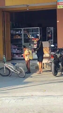 A man in a black long-sleeved shirt and shorts stands next to a child wearing a brown uniform and a yellow backpack. The man appears to be adjusting or handing something to the child, possibly within the backpack. They are standing outside an open storefront, which has shelves displaying various items. A white bicycle is parked to the left of the child, and a motorcycle is parked to the right. The ground is paved, and shadows indicate it is daytime.