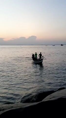 The video shows three people in a small, green boat on the water. They appear to be using oars. The water is calm and the sky is a pale white, suggesting either early morning or late afternoon. Other boats are visible in the distance. Large, dark rocks are in the foreground, likely part of a shoreline.