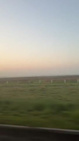 The view from a vehicle, likely a car or bus, moving along a highway at what appears to be dawn or dusk. The sky is a pale blue, tinged with a soft pink or orange near the horizon. A flat expanse of land stretches out beyond the highway. The land is a mix of green grass and brown fields. The foreground is dominated by the metal guardrail of the highway, which is blurred due to the motion of the vehicle. The overall impression is of a peaceful, rural scene at the edge of day or night.