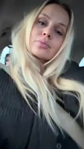 A woman with long blonde hair is seated in a car, wearing a black jacket and a seatbelt. She appears to be looking at the camera, running her fingers through her hair, and talking.