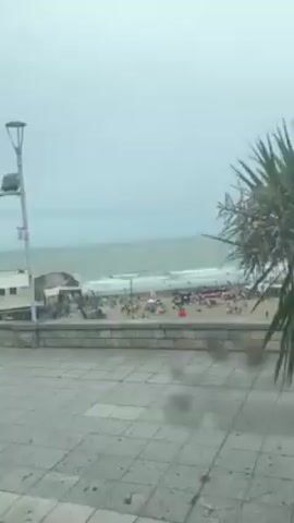 The camera is filming from inside a vehicle passing a beach on a cloudy day. The beach is crowded with people and a few umbrellas. In the background, the ocean waves are visible. A white building is situated next to the beach. A street lamp and a street sign are also visible. The ground is paved with grey tiles.