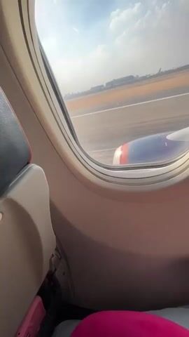 The video shows a view from an airplane window as it taxis on a runway. The camera is positioned inside the cabin, looking out. The window frame is visible, with part of a seat on the left. Outside the window, the runway stretches out, with markings and other airport structures in the distance. An airplane engine is visible to the right of the frame, with its blue and orange casing prominent. The sky is partly cloudy. The plane appears to be moving slowly.