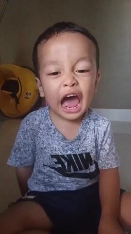 A young boy with short black hair and a grey t-shirt with a Nike logo is sitting down, looking at the camera and smiling. He sticks his tongue out at the camera in some frames. A yellow object is visible behind him on the left. The background appears to be a wall and a bathtub.