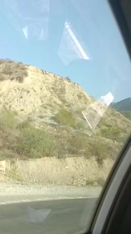 The video shows a view from a vehicle traveling along a road. The road appears to be paved with asphalt and gravel on the side. A dry, rocky hillside with sparse vegetation is visible on the right side of the frame, sloping upwards. In the distance, mountains with a similar dry, rocky terrain can be seen under a clear blue sky. There are reflections on the window, obscuring some parts of the view. A few white pieces of paper are visible on the road surface. Towards the end of the clip, a person's hand and arm are reflected in the window, making it appear as if they are interacting with the scenery. There is also a sign on the side of the road with red and white chevrons, indicating a curve or hazard.