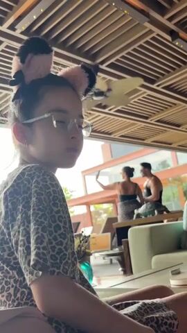 A young girl wearing glasses and a leopard print shirt with a fluffy headband sits in the foreground, looking down and to the right. Behind her, a woman in a black top and leopard print skirt is visible, as is a man in a black tank top. They are standing near a window or glass wall, with lush greenery and trees visible outside. The interior of the room has a distinctive wooden slatted ceiling and what appears to be a ceiling fan. The overall atmosphere suggests an indoor dining or lounge area, possibly at a resort or restaurant.