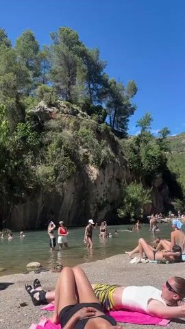 The video shows a beach with people swimming, sunbathing, and relaxing on a sunny day. A group of people in bathing suits are in the water, some standing and some swimming. On the sandy beach, several people are lying on towels, sunbathing. Others are sitting and talking. A person in a white shirt stands holding a reflector screen. The scene is surrounded by green trees and a clear blue sky with a few clouds. There is a mountain in the background, also covered with trees.