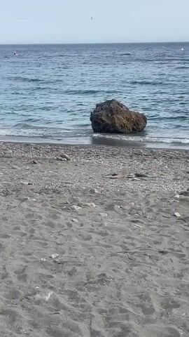 The video shows a rocky beach with dark sand in the foreground. The sea is visible in the background, with a large rock partially submerged in the water. Small waves are breaking on the shore. The sky is clear and pale blue. There is a small, dark shape in the distance on the water, possibly a person or a boat. The video appears to be shot from a stationary position on the beach, looking out towards the sea.