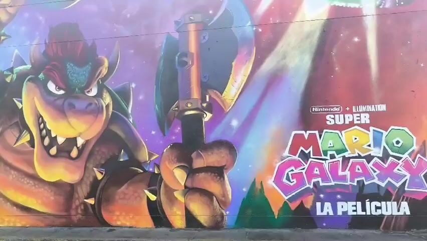 The video shows a mural on a wall advertising "Super Mario Galaxy: La Pelicula" (The Movie). The mural features Bowser holding an axe, along with other characters and elements from the Super Mario Galaxy game against a background of space. The mural stretches across a long wall next to a road.