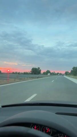 A first-person perspective video from inside a car shows a highway at dusk. The sky is cloudy with streaks of pink and orange from the setting sun. Trees and a guardrail line the left side of the road. Several trucks and cars are visible in the distance on the highway. The dashboard and steering wheel of the car are partially visible in the foreground. The road appears to be a multi-lane highway.