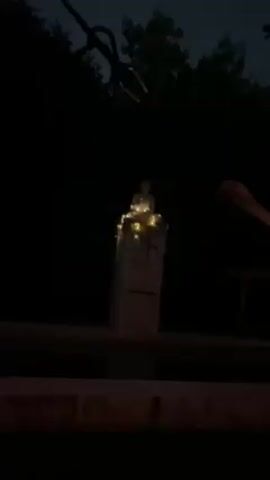 In the darkness of night, the camera focuses on a small, seated statue atop a light-colored pillar. The statue is adorned with small, warm-toned fairy lights, which provide the primary illumination, making the statue and pillar visible. The surrounding environment is dark, with glimpses of trees and possibly power lines or branches silhouetted against the night sky. To the right, there is the suggestion of a structure or object partially visible in the darkness. The shot is steady, with no apparent movement in the camera's position. The scene exudes a serene and tranquil atmosphere, highlighted by the soft glow of the fairy lights.