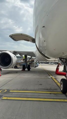 The video shows the back section of a large white passenger airplane, its cargo door open. Workers are present around a cargo lift and a fuel truck refuelling the plane. The plane is stationary on a concrete tarmac, marked with yellow lines and numbers. The weather is overcast, with a cloudy sky. Other airplanes are visible in the background, including one with the flag of Israel on its tail. The overall scene shows routine airport maintenance and preparation for flight.