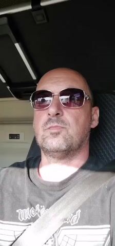 The video shows a close-up, head-and-shoulders shot of a bald man wearing sunglasses and a dark t-shirt with a light-colored seatbelt across his chest. The man has stubble and is looking slightly off-camera to his left. The t-shirt has gothic-style lettering visible on it, with the word "motor" clearly discernible. The background suggests an indoor setting, possibly the interior of a vehicle, with some lighting fixtures visible overhead and a grey, textured seat behind the man. The lighting indicates it is daytime. The man's expression appears neutral or slightly serious. There are no other people or objects prominently featured in the frame. The location context provided is Mogila, Bulgaria, but there are no specific visual cues in the video to confirm this.