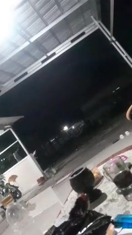 The video shows a dark, outdoor scene at night. In the foreground, on a table covered with a lace tablecloth, are several objects. These include a glass with a dark liquid, a small bowl with some food items, a dark-colored decorative object, and a cigarette pack with a blue lighter. A portion of a person's arm and shoulder, wearing a dark garment, is visible on the right side of the frame. In the background, there is a metal roof structure, and beyond that, a dark open space with some faint lights, possibly from a street or distant buildings. The overall impression is that of a casual, possibly late-night gathering or a moment of relaxation outdoors.