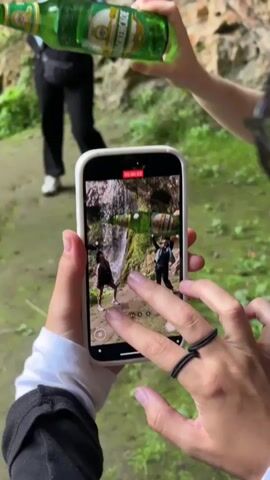 A person holds a smartphone in portrait mode, recording a video. The phone screen shows a scene with two individuals standing near a waterfall. One individual, on the left, poses with hands on hips. The other, on the right, raises their arms and holds a green bottle of liquid above their head. A third person, out of focus in the background, walks away from the camera, holding a similar green bottle. The environment appears to be a rocky area with lush green foliage and a waterfall cascading down rocks. Another person's hands are visible in the foreground, their fingers interacting with the smartphone screen. The timestamp on the phone indicates the video is just a few seconds long.