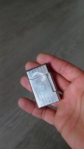 A hand holds a silver lighter with an engraved pattern. The lighter is closed at the beginning of the video. The hand turns the lighter, showing different angles. In the last few seconds, the hand opens the lighter, revealing the flame mechanism. The background appears to be a wooden floor.