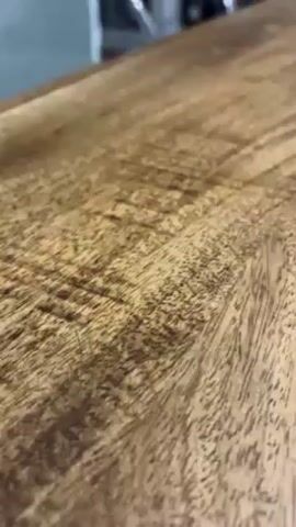 The camera is focused on a wooden countertop, likely a kitchen island, with a visible wood grain pattern. In the blurred background, there is a white refrigerator, black appliances, a light-colored chair, and a brown dresser. The countertop is the main element in focus, showing its texture and color. The video is static and doesn't reveal any movement or interactions.