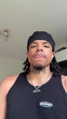 A man wearing a black durag, a black tank top, and white earbuds is looking at the camera. He has braids that extend past his shoulders and a tattoo on his neck. He is wearing a silver chain with a pendant. The background appears to be a kitchen with a refrigerator, white cabinets, and a stove top with pots and pans. The lighting suggests it is daytime. The man is likely recording a video or taking a selfie.