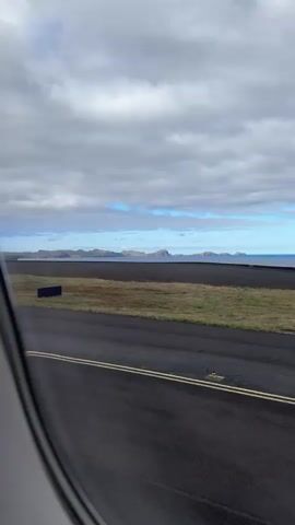 The video shows a view from inside an airplane, looking out of a window at an airport runway. The runway is black asphalt with yellow lines. On either side of the runway is grass. Beyond the runway is the ocean, and in the distance are islands under a cloudy sky. The video appears to be from the start of a taxi.