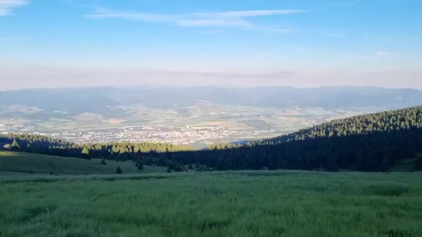 The media file shows a panoramic view of a valley and distant mountains, likely taken from a hilltop or mountain slope. In the foreground, there's a field of tall green grass, with a line of dark green coniferous trees forming a dense forest along the mid-ground to the right. Beyond the forest, a town or city is visible, characterized by its buildings and structures spread across the valley floor. The landscape stretches out to a series of undulating hills and mountains in the far distance, which appear hazy due to atmospheric perspective, suggesting a significant distance.

The sky is a clear blue with a few wispy clouds, indicative of daytime. The lighting suggests it's either early morning or late afternoon, with a soft, warm light illuminating parts of the scene. The shadows cast by the trees indicate the direction of the sun. The overall mood of the scene is serene and peaceful, conveying a sense of natural beauty and vastness. The location is identified as Martin, Slovakia, which provides context for the mountainous terrain and the presence of a town nestled in the valley. There is no visible text within the media file.