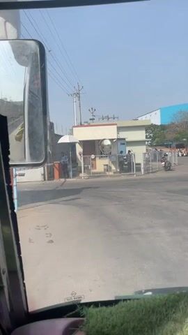 The video shows a view from inside a bus driving through a parking lot. In the first shot, the bus is near a gated entrance with a security booth. A person is visible inside the booth, and motorcycles and a small truck are parked nearby. As the bus moves forward, a view of the parking lot comes into focus, showing several parked trucks and buses, as well as people walking around. The bus passes a yellow bus parked in front of it, and a traffic cone can be seen on the road.  In the background, there are trees and what appears to be the edge of a building with a blue roof.  The video is shot during the day with clear skies.
