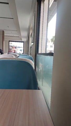 The video provides a low-angle, close-up view of a restaurant or cafe interior. The foreground is dominated by a wooden table surface. Beyond the table, a row of blue upholstered chairs with white patterned cushions is visible, arranged along the side of the establishment. A large window runs along the length of the chairs, offering a view of the outside. Through the window, a street scene can be seen, including buildings, trees, and pedestrians. The overall environment suggests a public dining or waiting area, possibly during the daytime given the natural light filtering through the window. There are no explicit actions or interactions captured in the brief clip, focusing instead on the static elements of the setting.