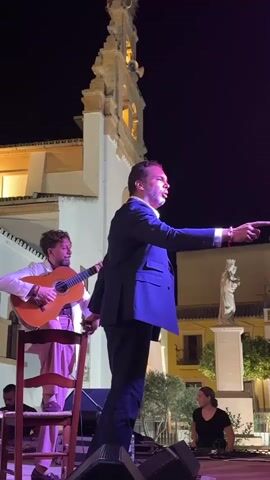 A man in a dark suit stands on a stage, pointing with his right hand. A person playing a guitar sits on a chair to his left. A woman is seated on the stage to the right of the man in the suit. The background shows a building with a broken tower and a statue. The scene is illuminated by stage lights.
