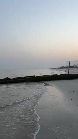The video captures a serene beach scene at what appears to be dawn or dusk. The sky is a soft gradient of pale blue and pink, hinting at the transition of light. The ocean water is calm, with gentle waves lapping at the sandy shore. In the foreground, the water is a murky gray-green, and small whitecaps indicate the movement of the waves. The sand on the beach is a light tan color, with a damp sheen where the waves have recently receded.

To the left of the frame, a structure made of large, rough-hewn concrete blocks, covered in dark green moss, juts out into the water. These blocks form a breakwater or some kind of man-made barrier against the sea. The structure is partially submerged, with waves washing over the tops of some of the blocks.

Further down the beach, towards the right side of the frame, a tall, square metal fence structure stands on the sand. It is made of dark metal poles and wire mesh, and it extends vertically upwards, appearing to be a protective enclosure or boundary marker. The background shows a hazy horizon where the sea meets the sky, with faint, indistinct shapes that could be distant buildings or landforms. The overall atmosphere is tranquil and natural, with a hint of human intervention in the form of the concrete structure and the fence. There are no people visible in the video.