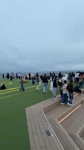 The video shows a group of people on a rooftop gathering area. The area has tiered wooden seating, artificial turf, and a glass structure. People are standing and socializing, with some looking out at the city view as the sun sets on the horizon.  The scene suggests a casual event or gathering taking place at an elevated location, potentially a rooftop bar or observation deck.