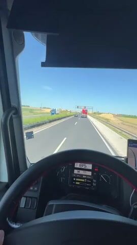The video is filmed from the perspective of a truck driver on a highway. The dashboard of the truck is visible in the foreground, displaying a speed of 85 km/h. A smartphone mounted on the dashboard shows a navigation app. The road ahead is clear, with other trucks in the distance. The environment appears to be rural, with fields on either side of the highway under a clear blue sky. The driver's hand is seen briefly on the steering wheel.