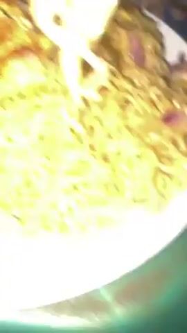 A fork scoops up noodles from a white plate piled with cooked noodles and a large onion omelet. The person holding the plate is sitting in a blue chair.