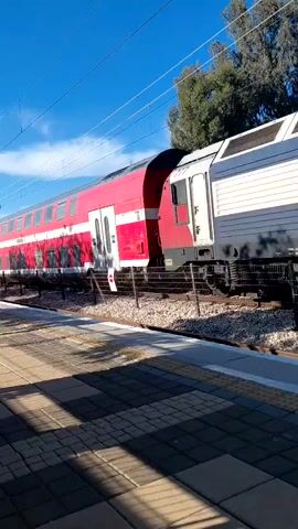 A red and white double-decker train passes a train platform. The train is in motion, and the camera follows it from a stationary position on the platform. The sky is blue with some clouds. There are power lines and a tree in the background. The platform is made of gray rectangular pavers. A black fence separates the platform from the train tracks.