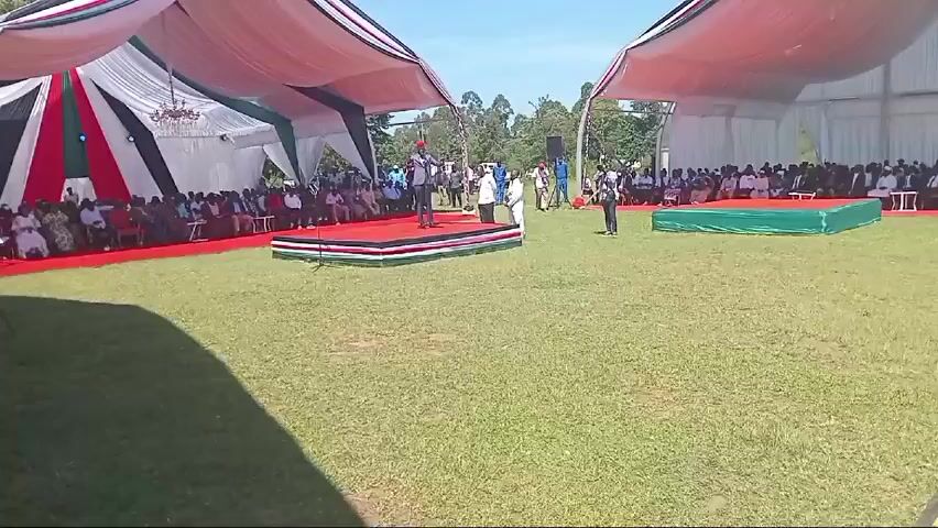 The video takes place outdoors, where a man wearing a red hat and a grey suit is speaking at a microphone on a stage covered in red carpet. The trim on the stage is black, white, red, and green, which resembles the flag of Kenya. He appears to be giving a speech to a seated audience under tents. There are other men standing nearby and a cameraman is present.