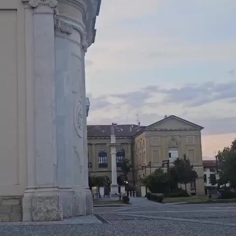 The video displays a slow zoom-out from a close-up of a white stone pillar to reveal a town square. In the foreground, the left side of the video is dominated by the large, ornate white stone pillar of a building. The pillar is textured and features decorative carvings. The ground in front of it is paved with small, dark gray stones.

As the camera zooms out, a central monument comes into view. This monument appears to be a tall column topped with a statue of a figure, possibly religious, with a halo. Behind the monument are several buildings. The main building in the center is large and appears to be a historical structure with a tiled roof and multiple windows, some of which are arched. To the right of this building is another large, light-colored building with a more classical architectural style, also featuring numerous windows. Trees and greenery are visible in front of these buildings.

The sky above is overcast with scattered clouds, and the light suggests either dawn or dusk. The overall impression is of a quiet, historic town square. No people or animals are visible in the video.
