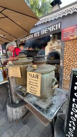 The video shows an outdoor food stall with the sign "Пирожки из печки" (Pies from the oven). In the foreground, there are two large, antique samovars, one on each side of the frame. Both samovars are steaming, indicating they are in use. The samovar on the left has a sign that reads "Сбитень" (Sbiten), and the one on the right has a sign that says "Чай" (Tea), listing ingredients such as Ivan tea, mint, lavender, and ginger. Hanging from the samovar on the left are several small, dried bread rings, or baranki. A man wearing a red shirt and a cap is visible behind a table on the left, appearing to be working at the stall. Behind the samovars, a brick oven is visible with flames inside, suggesting that pies are indeed being baked there. Wood logs are stacked to the right of the oven. A woman is partially visible on the right side of the frame, appearing to interact with the samovars or the stall. The overall scene suggests a traditional Russian food vendor.