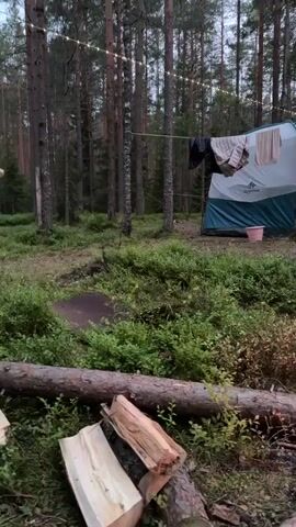 The video shows a campsite in a forest. In the foreground, there is a campfire with logs burning, and several cut logs are placed nearby. Behind the campfire, there is a green tent with clothes hanging on a line in front of it. A white and green striped camping chair is visible to the right of the tent. Further back, there are more tents and tarps, as well as some people. One person is sitting in a chair in front of the campfire, wearing a red jacket and black pants. Another person is partially visible in the foreground, seated on the right. The environment is a forest with tall trees and undergrowth. The lighting suggests it is dusk or early evening.