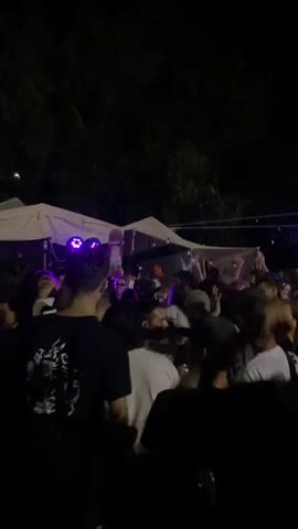 This is a vertical, full-frame, shaky video taken at night from behind a crowd of people. The crowd is facing a stage area where there are white tents. Blue and purple lights are visible, as are what appear to be speakers and a skateboard. People in the crowd are dressed casually. The overall atmosphere appears to be a nighttime outdoor event or party.