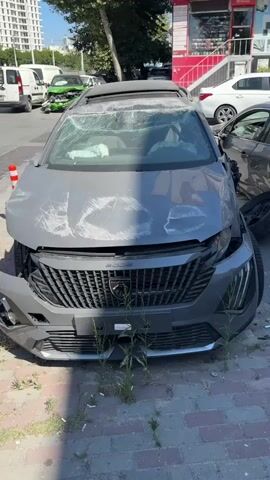 The video shows a grey Peugeot 3008 that appears to have been in a serious accident. The windshield is heavily cracked, and the hood is also damaged with visible scratches. The front bumper is detached on one side, and the car's front left fender is crumpled inwards. The front airbags appear to have deployed, indicated by white fabric visible through the cracked windshield. The car is parked on a paved street, with vegetation growing around its front. In the background, other vehicles and buildings are visible, suggesting an urban or suburban environment. One of the background vehicles, a green car, also appears to be damaged. The overall scene suggests the aftermath of a collision.